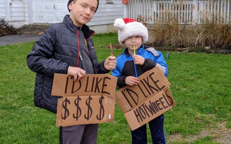 Two children holding cardboard signs that tell Santa what they want for Christmas: I'd like $$$ and I'd like hotwheels.